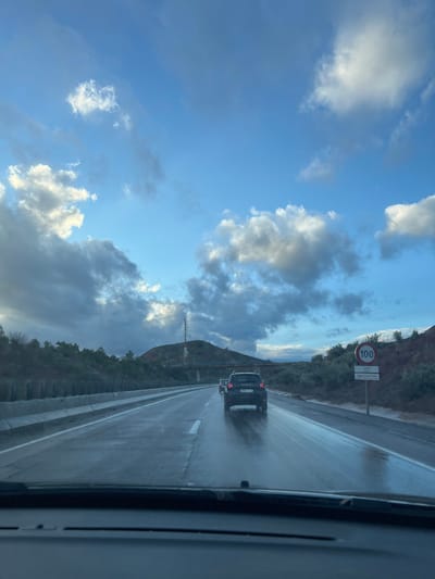 A road with a car in the distance under a cloudy sky. The road appears wet, possibly from rain. A 100 km/h speed limit sign is visible on the right side. The landscape includes a mountain range partially obscured by clouds. The photo seems to have been taken from inside a moving vehicle, with a portion of the vehicle's interior visible on the right. The sky is blue with white and grey clouds.
