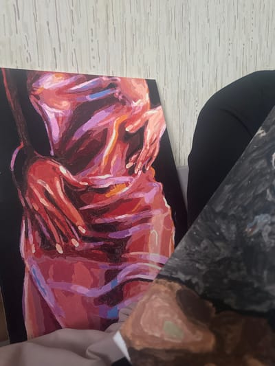 The image shows a painting lying on a surface that appears to be light-colored wood grain. The painting features two figures in a stylized, somewhat abstract manner, rendered in shades of red, pink, purple, and brown. The figures are intertwined and appear to be lying down, with hands prominently displayed. The background of the painting is black. Parts of other objects are present in the image, including an expanse of dark fabric and a section with shades of gray, brown, and white. The image does not provide enough information to determine the location or any details regarding the city of Высокий, Russia.