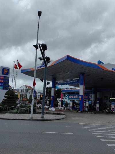 The image depicts a Petrolimex gas station situated at a street corner under an overcast sky in Thuận Hóa Ward, Vietnam.
The Petrolimex station features a large canopy with a prominent blue and orange trim, supported by several blue pillars. A white sign on the canopy identifies it as "CÔNG TY XĂNG DẦU THỪA THIÊN HUẾ PETROLIMEX-CỬA HÀNG 01" (Thua Thien Hue Petroleum Company Petrolimex - Store 01). To the left, a tall sign displays the Petrolimex logo and digital prices for various fuel types, including "95-V," "95-IV," and "DO-V." Two flagpoles stand near this sign, flying the red Vietnamese national flag with a yellow star and a solid red flag. Under the canopy, multiple fuel pumps are visible, alongside several parked motorbikes and a few individuals; one person appears to be fueling a motorbike. Behind the pumps, part of a building is visible with "KÍNH MẮT" (eyeglasses) signage.
In the foreground, an asphalt road includes a solid white line and a designated pedestrian crossing with white zebra stripes. A concrete sidewalk curves around a small, fenced, triangular median strip that contains a manicured, tiered green bush. Two tall poles stand at the intersection: one a street lamp, the other supporting multiple clusters of traffic lights. A small red flag is attached to one of these poles. In the background, across the street from the gas station, various multi-story buildings and additional vehicles are visible. The sky is uniformly gray and overcast.