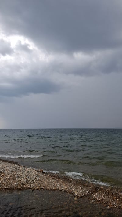The image depicts a moody, overcast scene at the shore of a large body of water, possibly Lake Baikal, near the city of Priboi, Russia. The sky is filled with heavy, grey clouds, suggesting an impending storm or recent rainfall. The water is a deep, dark blue-green, with visible ripples and small waves lapping against the shore. The shoreline is composed of smooth, multicolored pebbles and stones, with a narrow strip of darker, wet sand or mud closer to the water's edge. The overall atmosphere is somber and natural, with no signs of human presence or activity.