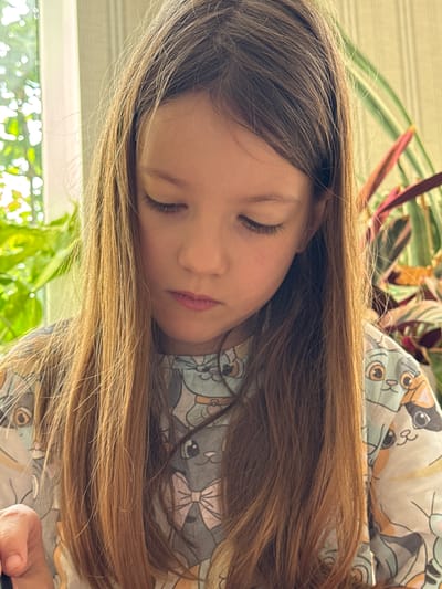 The image shows a close-up, side profile of a young girl sleeping. She has long, light brown hair that falls around her face and shoulders. Her eyes are closed, and her lips are slightly parted. She is wearing a light-colored shirt with a repeating pattern of cartoon cats. The background is a blur of green foliage and what appears to be a textured wall or blinds. The lighting suggests it is daytime, possibly indoors near a window given the bright, diffused light. The overall mood is peaceful and serene, conveying a sense of rest. The image is oriented vertically, with the girl's head on the left and her body extending to the right. No text is visible in the image.
