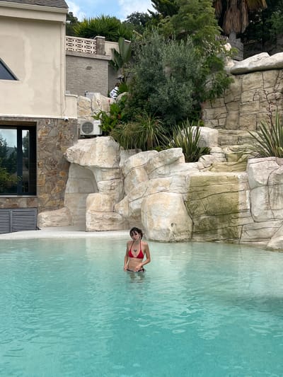 In the city of Can Güell, Spain, a young woman with dark hair, wearing a red bikini, stands in a clear turquoise swimming pool. She is partially submerged, with the water reaching her waist. Her arms are by her sides, and she is looking towards the camera. The pool is surrounded by a large, artificial rock formation that resembles a waterfall. Behind the rock formation, there is a building with a textured stone facade and a dark-framed window. Further back, lush green foliage and trees are visible against a cloudy sky. An outdoor air conditioning unit is mounted on the rock face.