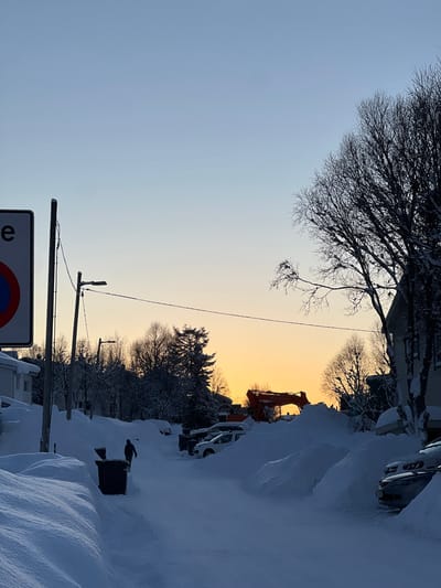 A winter landscape shows snow-covered streets with parked cars buried in drifts. An orange-hued sky appears between snow-laden trees. Power lines stretch across the sky, connecting poles along the street. A construction vehicle, possibly an excavator, is visible amidst the snowdrifts, suggesting snow removal operations. A street sign stands in the foreground.
