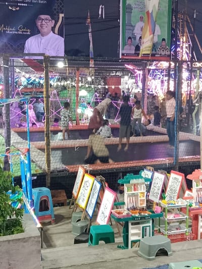 The image shows a trampoline enclosed by a black metal fence. There are six people, including one adult man, and five children, all wearing casual clothing. Some of the children are jumping on the trampoline, while others are standing on the sidelines. The trampoline is located in a night market or fairground, and the surrounding area is brightly lit with string lights and other decorations. The image was captured in Kaliwungu, Indonesia, at coordinates -6.962, 110.253. There are also two toy kitchens, small easels, and small plastic chairs on the ground near the fence. A large poster advertising a local candidate for the DPR (Indonesian House of Representatives) can be seen in the background. The poster reads "Mahfud Goding, Kandidat DPR Kob, Kendal." The scene appears to be lively and festive.