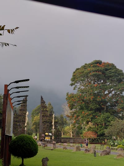 The image depicts the Pura Ulun Danu Batur temple in Sukasada, Indonesia, under an overcast sky. In the foreground to the left, a wooden structure with multiple modern streetlights is visible. To the right of this, a dense, spherical green shrub stands out against a well-manicured lawn. In the mid-ground, two imposing stone temple gates, intricately carved and adorned with decorative elements, stand sentinel. Beyond the gates, lush green trees and foliage dominate the landscape, with a large tree bearing vibrant orange blossoms taking center stage. A few people are visible walking in the distance, one couple strolling on a pathway and another person standing near the temple gates. The grounds are covered in grass, interspersed with large, rounded stones that appear to be decorative. The background is shrouded in a soft mist, obscuring the distant hills and mountains.