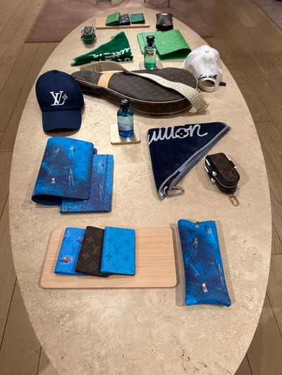 The image depicts an overhead view of an assortment of Louis Vuitton merchandise arranged on an oval-shaped light-colored stone-like surface.

Items present include Louis Vuitton branded towels, caps, small bags, sandals, notebooks or folders, and what appears to be a perfume bottle. Some items bear the classic LV monogram pattern. A wooden board supports some of the notebooks or folders.

Given the presence of high-end merchandise, the location is likely a retail environment. The city mentioned, Oak Brook, United States of America, suggests that this might be the Louis Vuitton store.