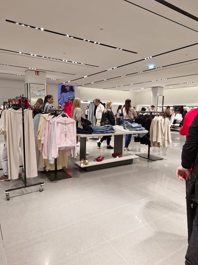 This image depicts the interior of a clothing store, likely part of a chain like Zara, given the style of clothing and display. The environment is modern with a bright, tiled floor and a suspended ceiling with track lighting. Several individuals, predominantly women, are browsing the merchandise. Clothing racks display various garments, including casual wear like t-shirts, hoodies, and pants, as well as more formal items like blazers. One display table holds folded clothing and a pair of red high-heeled shoes. The store appears to be well-lit and organized, with a significant amount of inventory visible. The presence of a large screen displaying a fashion model suggests a contemporary retail setting. The photograph was taken in Bremen, Germany.