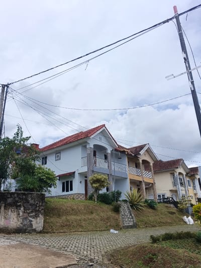 A row of two-story houses with red tile roofs sits on a grassy hill. The houses vary in color, including white, beige, and light brown. Each house has a balcony or covered porch with columns. Power lines crisscross the sky above, anchored by utility poles. Paved pathways lead up to the houses. A stone wall sits in the foreground, bordering the street, with some foliage growing. The sky is cloudy and overcast.