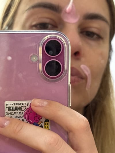 An indoor mirror selfie captures a person engaged in a skincare routine. The subject's face is visible in reflection, partially out of focus, showing one eye, an eyebrow, and parts of her nose and mouth. She has streaks of a light pink substance, likely a face mask or cream, applied to her forehead and nose area. Her light-colored hair is visible on the left side of the reflection.
The person is holding a light purple smartphone, encased in a clear protective cover, which dominates the foreground. The phone features two prominent circular camera lenses and a smaller sensor below them. Her right hand, with manicured nails (clear polish), holds the device. A sticker, partially obscured by her fingers, is affixed to the back of the phone. The text on the sticker, reversed due to the mirror reflection, reads in Russian: "Холодная СНИМКОЦ" (Kholodnaya SNIMKOTs), meaning "Cold pictures," and a sequence of numbers and letters "97892979 В".
The image portrays a personal moment of self-care, possibly taken in a bathroom or bedroom in Ust-Ilimsk, Russia. The time of day and specific weather conditions are not discernible from the image.