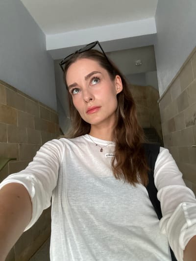 A woman is standing in what appears to be a bathroom with light-colored, possibly tiled walls and flooring. She is wearing a white long-sleeved shirt. Part of her hair is light brown and wavy. She is looking slightly upwards and to the left. Glasses are visible on her head. Her facial features are discernible, with fair skin and makeup on her eyes and lips. A necklace is visible around her neck. The location of this image cannot be determined to be in Kiel, Germany.