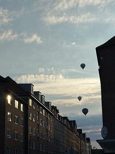 The image captures a street scene in Kiel, Germany, under a partially cloudy sky. Several hot air balloons are visible in the distance, floating above a row of brick buildings. The buildings have a consistent architectural style, featuring multiple stories, numerous windows, and some dormer windows on the rooftops. The sun is setting or rising, casting a warm glow and creating reflections on some of the building windows, particularly on the left side of the image. The overall impression is a serene urban landscape with a touch of whimsy due to the presence of the balloons.