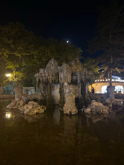 The image depicts a nighttime scene featuring a large, rocky fountain with stalactite-like formations, situated in a body of water. The fountain is surrounded by rocks and vegetation, with trees visible in the background. A streetlamp illuminates the left side of the scene. In the distance, a building with arched windows is lit up, and a person is silhouetted against the light. The sky is dark, with a sliver of the moon visible. This scene is located in the city of Pyatigorsk, Russia, specifically within the grounds of the "Lermontov Gallery," a cultural and historical site. The overall impression is one of a serene, somewhat mysterious, nocturnal ambiance.