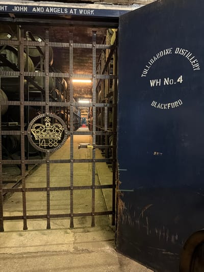 The image shows the entrance to a whisky warehouse. A sign above the entrance reads "Tullibardine Distillery, WH No. 4, Blackford." A metal gate with a central circular emblem of a crown and the number 1488 guards the entrance. Barrels are visible inside the warehouse, stacked on racks. A sign to the left of the gate reads "RIGHT JOHN AND ANGELS AT WORK." The surrounding structure appears to be made of concrete. The location is Blackford, United Kingdom.