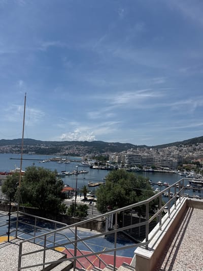 This image captures a panoramic view of the city of Kavala, Greece, featuring a harbor filled with numerous boats and a cityscape climbing the hills. In the foreground, a portion of an outdoor basketball court with blue and red surfaces is visible, bordered by a gray railing and textured stairs. Beyond the court, lush green trees partially obscure the view of the harbor. The harbor itself is a hub of activity, with a dense collection of various boats, including sailboats and motor yachts, docked in the clear blue water. A long, slender pole with a cross at its apex stands prominently in the left side of the frame. The backdrop is a sprawling urban landscape of white and light-colored buildings that extend up the gentle slopes of the surrounding hills, dotted with more greenery. The sky above is a vibrant blue, streaked with wispy white clouds, indicating fair weather. The overall scene suggests a sunny day in a coastal Mediterranean city.