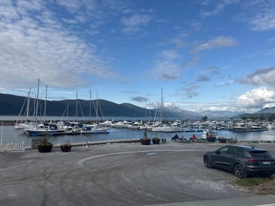 This media file depicts a scenic view of a marina in Nesna, Norway. The sky is a vibrant blue, dotted with fluffy white clouds, suggesting pleasant weather, possibly late afternoon. The marina is filled with numerous sailboats and motorboats docked in the calm, dark blue water. In the background, majestic, verdant mountains rise, some capped with snow, adding to the dramatic natural beauty of the location.

In the foreground, a gravel area leads to the marina. A dark gray SUV is parked on the right, with a license plate clearly visible reading "EP 63900". To the left of the SUV, two large planters with small trees and plants add a touch of greenery. Further left, a couple of people are seated at a picnic table, seemingly enjoying the view. Another person is seated on a bench. The overall atmosphere is peaceful and serene, indicative of a relaxed day at the harbor. The presence of boats and the mountainous backdrop strongly suggest a coastal or fjord setting.