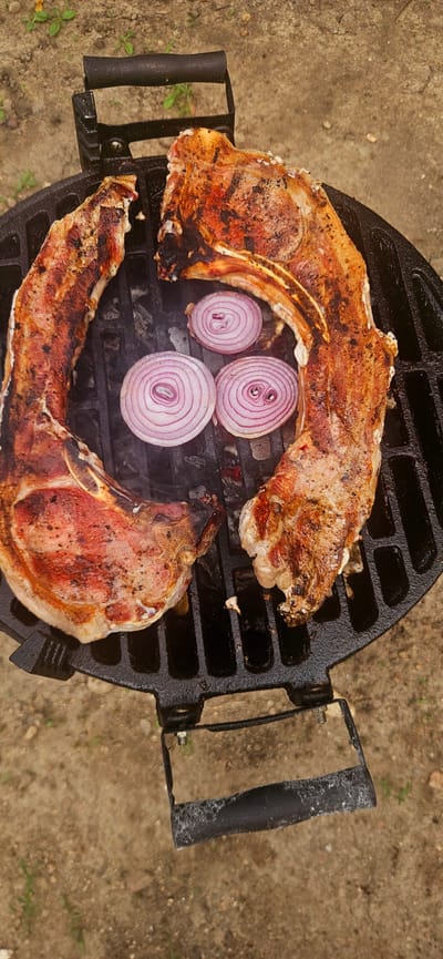 Two large, bone-in pork chops are being grilled on a black charcoal grill. The chops are partially cooked, with visible grill marks and browning. Three rings of red onion are placed on the grill next to the meat. The grill is outdoors, with a dirt and gravel ground surface visible. The background shows sparse green vegetation. The scene is photographed from a top-down perspective. The location is the city of Kostenets, Bulgaria.