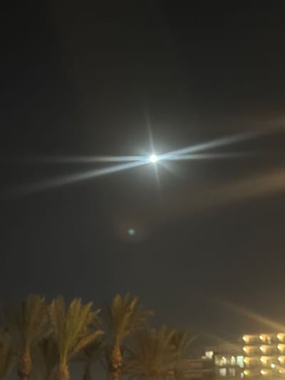 The image shows a night scene in Saint Paul's Bay, Malta. A bright light source, possibly the moon, is visible in the upper center of the frame with distinct light rays emanating from it. To the right, out of focus, is the top of a possible structure or decoration. The sky is dark, and the overall atmosphere is hazy.