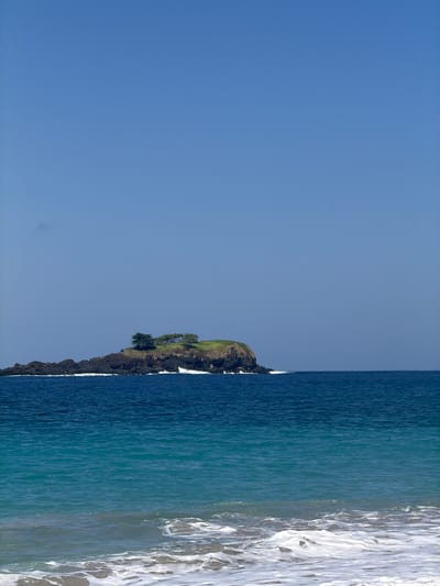 A rocky island with a grassy top and a few trees stands in the ocean off the coast of Karangasem, Indonesia. The water is a deep blue, transitioning to a lighter turquoise closer to the shore. Waves crash against the rocks, creating white foam. The sky is a clear, bright blue.