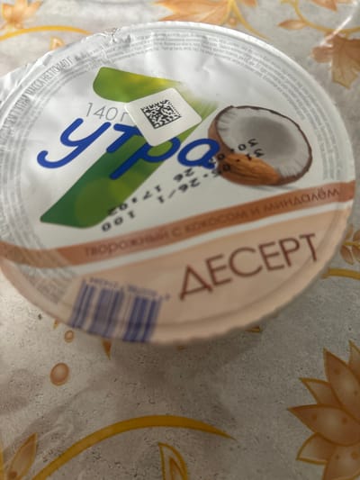 The image shows a round, plastic container of yogurt dessert. The container has a lid with a picture of a coconut half and an almond, along with text in Russian. The text on the lid indicates it is a "Dessert with coconut and almond" and includes nutritional information such as fat content (3%) and weight (140g). There is also a QR code on the side of the container. The container is placed on a patterned tablecloth, possibly in a home setting. The lighting suggests it is daytime, and there are no indications of specific weather conditions. The image appears to be a product shot or a casual photo of the food item. The text is primarily in Russian, with some numbers and symbols.