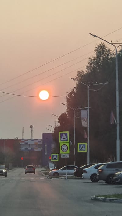 The image captures a street scene in Megion, Russia, during sunset. The sun, a bright, hazy orb, hangs low in the sky, casting a warm, orange glow over the surroundings. Silhouetted against the sunset are trees and power lines, contributing to a somewhat hazy atmosphere, possibly due to dust or smoke.
The scene features a road with vehicles, including a silver car parked on the right and a darker car further down the road. Several traffic signs are visible, including pedestrian crossing signs, a speed limit sign indicating 20, and a sign with "3.5 M". The presence of these signs and the road infrastructure suggests an urban or suburban setting. In the background, parts of buildings and what appear to be construction cranes can be seen, indicating ongoing development or industrial activity. The overall mood is serene, with the warm colors of the sunset creating a peaceful ambiance. The time of day is clearly evening, as indicated by the low position of the sun and the soft lighting.