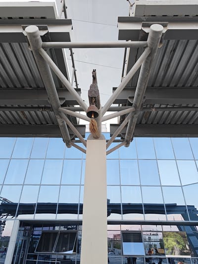 This media file features an upward-angle shot of a building's exterior, focusing on architectural elements and reflections. The primary subjects are the modern building's facade, composed of large, reflective glass panels, and a supporting structural framework. The glass reflects the sky and surrounding environment, creating a dynamic visual effect.

Attached to the structural beams is a decorative element consisting of a bell and a tassel, possibly an artistic or cultural accent. The metal framework is industrial in appearance, with visible pipes and beams, painted in a light color. Below the main structure, the lower section of the building shows windows, some open, and reflections of buildings and trees. A few people are visible as blurry figures in the reflections, suggesting an outdoor setting.

The time of day appears to be late afternoon or early evening, as indicated by the soft lighting and the reflections showing a mix of bright sky and warmer tones from the buildings. The weather seems clear, with a bright blue sky reflected in the glass. The location is implied to be Yakutsk, Russia, based on the contextual information. No specific activity or event is clearly depicted; the focus is on the architecture and the interplay of light and reflection. There is no discernible text in the image.