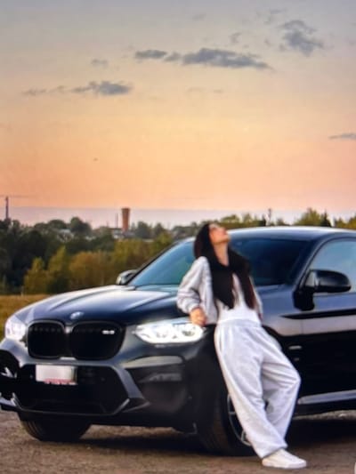 A full-body shot features a young woman leaning against a black BMW SUV. The woman, with long dark hair, wears a light-colored tracksuit and white sneakers. She is looking upwards, with her head tilted back. The SUV is parked on a dirt or gravel surface. In the background, a line of trees and distant industrial structures are visible against a sky with soft clouds, colored by sunset. The overall scene suggests an outdoor setting, possibly on the outskirts of a town or in a rural area, given the presence of industrial elements and a more natural landscape. The lighting indicates it is late afternoon or early evening. The specific location is Beșalma, Moldova, as indicated by the metadata.