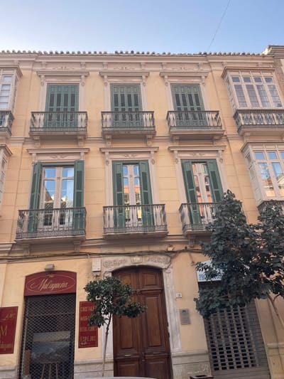 The image shows a building facade in Málaga, Spain, under a clear sky. The building has a light yellow stucco exterior with ornate detailing around the windows and roofline. There are multiple stories with several windows on each level, many featuring green shutters and small balconies with decorative iron railings. Some of the windows are open, revealing reflections of buildings and the sky.

On the ground floor, there are storefronts. The left storefront has a red sign with the word "Malagueña" in an elegant script, and the storefront itself is covered by a metal security gate. To the right of this, there is a large wooden door. Adjacent to the door, there is another storefront with a blue sign that appears to say "PROSPER," also covered by a metal security gate. A small tree with dark green leaves is situated in front of the storefronts, partially obscuring the view of the ground floor details. The overall impression is of a historic building in an urban setting.