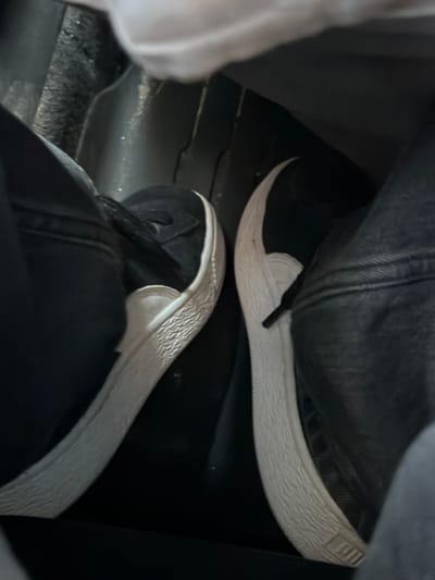 The image shows a close-up view of two feet wearing black sneakers with white soles. The feet are positioned inside what appears to be a vehicle, possibly in the footwell. The left shoe is closer to the foreground, and the right shoe is slightly behind and to the right. The environment appears to be dimly lit. There is no indication of the location in Kareby, Sweden.