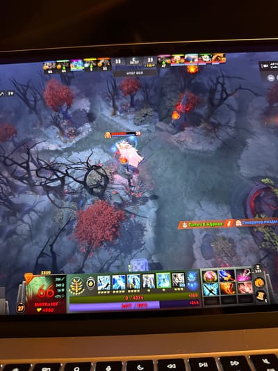 The image depicts a screenshot of a game of Dota 2 being played on a laptop. The game is in progress, with player avatars, health bars, and a minimap visible on the screen. The game's interface shows player statistics, item inventories, and chat messages. The environment within the game is a dark, ethereal forest with red-leaved trees and shadowy figures. The laptop is placed on a dark surface, likely a desk, with a portion of a keyboard visible in the foreground. The user's gaming setup appears to be in a dimly lit room, suggesting it might be nighttime or the room's lighting is subdued. There is no visible indication or context related to the city of Wervik, Belgium within the image. The scene is entirely digital and contained within the game's virtual world.