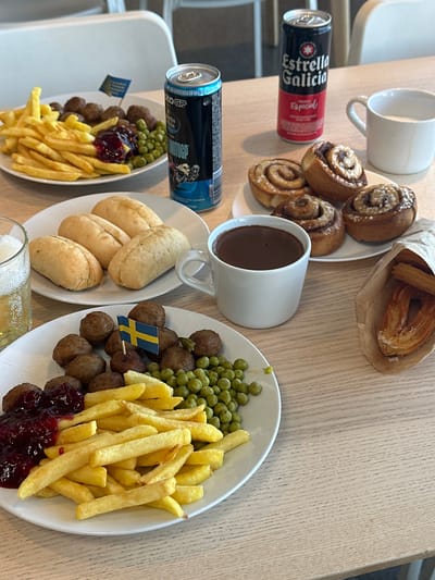 In a restaurant setting, a wooden table displays a meal consisting of Swedish meatballs, french fries, peas, and lingonberry jam. Accompanying the meal are two cans of Estrella Galicia beer, a mug of chocolate, and a bag of churros. In the background, white chairs and a glimpse of a window are visible. The presence of a small Swedish flag on a toothpick stuck into the meatballs suggests a thematic connection or restaurant. The lighting is natural and bright, illuminating the food and drinks on the table. The overall scene is a close-up shot of a culinary spread, with a specific focus on the food and beverages.