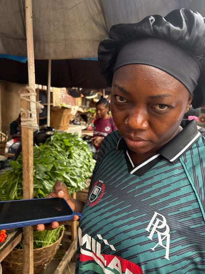 A medium-shot captures a woman holding a blue smartphone in a market setting, likely in Lugbe, Nigeria. She is turned at an angle towards the camera, wearing a black head covering and a green jersey with white lettering. Fresh greens are piled in front of her, suggesting she might be selling produce. In the background, other individuals and market stalls can be seen, contributing to the bustling atmosphere.