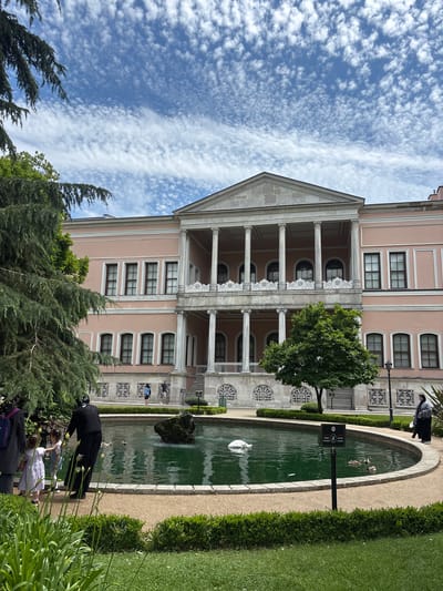 In the city of Beşiktaş, Turkey, an ornate, pink palace building stands under a partially cloudy blue sky. The palace features classical architecture with a colonnaded facade, balconies with decorative white railings, and numerous arched windows. In front of the palace, a circular pond with green water reflects the sky and surrounding greenery. A fountain is active in the pond, creating a spray of water. Several ducks swim in the pond, and a swan is also visible. A black rock formation is situated in the center of the pond. A sign with text and symbols is posted at the edge of the pond.

Several people are in the scene. In the foreground, to the left, a group of people, including two young girls in dresses and an adult in dark clothing, are interacting near the pond. To the right, near the pond's edge, a woman wearing a headscarf and a long dark dress stands. Further back, on the palace grounds, a few individuals are visible walking. Lush green trees and manicured hedges surround the pond and the palace, creating a park-like atmosphere. The ground around the pond is covered in light brown gravel.