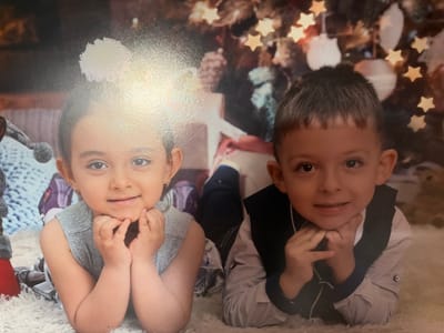 The image displays two young children, a girl and a boy, posed on a white, shaggy rug in what appears to be a photography studio. Both children are lying prone, facing forward, with their chins resting on their clasped hands, and both are smiling.

The girl, positioned on the left, has dark hair pulled back and is wearing a light-colored, sleeveless garment. Small earrings are visible. A significant bright glare obscures the upper left portion of the image, including part of the background and extending onto her forehead. To her immediate left, a red object resembling a boot and a grey knitted item with a pom-pom are partially visible, indicative of props.

The boy, positioned on the right, has short dark hair and is dressed in a light-colored, patterned long-sleeved shirt with a dark vest or jacket worn over it.

The background is a blurred photographic backdrop. On the right, elements of a decorated Christmas tree are discernible, featuring illuminated star-shaped lights and lighter-colored baubles. The left side of the background is more abstract, with soft, indistinct festive elements.

The scene contains no discernible landmarks, identifying text, or unique architectural features that visually confirm its location in the city of Kostinbrod, Bulgaria.