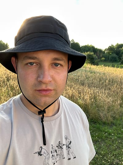 A medium shot captures a man from the chest up, wearing a black bucket hat and a light-colored t-shirt. The t-shirt has a graphic design of cartoon-like figures holding weapons. A black drawstring with a toggle closure hangs from the hat under the man's chin. The man's head is slightly tilted and he is looking directly at the camera. His eyes are open, and his expression is neutral.

The background consists of a field of dry grass illuminated by the setting sun, creating a golden hue. Beyond the field, a line of green trees forms a treeline against a bright, possibly hazy, sky. The lighting suggests it is late afternoon or early evening. The location is identified as Dubyshche, Ukraine.