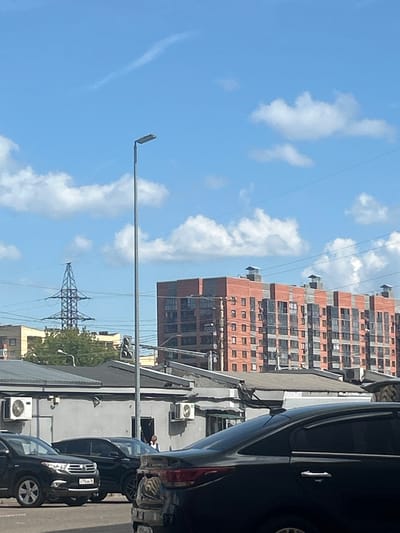 In Bor, Russia, a street scene unfolds under a partly cloudy blue sky. A tall, grey lamppost stands prominently in the foreground. Behind it, a cluster of grey, low-rise buildings with air conditioning units mounted on their facades are visible. Further in the background, a towering, multi-story residential building with red brick accents and numerous windows dominates the skyline. To the left, a power line tower with a lattice structure is visible. Several cars are parked and driving on the street, including black sedans and an SUV. A person, possibly male, is seen standing in an open doorway of one of the low-rise buildings. The overall impression is one of a typical urban environment on a clear day.