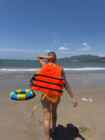 A person is standing on a sandy beach in Quy Nhon Ward, Vietnam, facing away from the camera and towards the ocean. The person is wearing an orange life vest and orange bikini bottoms. They have their left arm raised, with their hand behind their head. To the left of the person, a blue and yellow inflatable ring is lying on the sand. White flip-flops are visible on the sand to the right of the person. The ocean is visible in the background, with waves breaking on the shore. The sky is blue with a few scattered clouds. The overall impression is a sunny day at the beach.