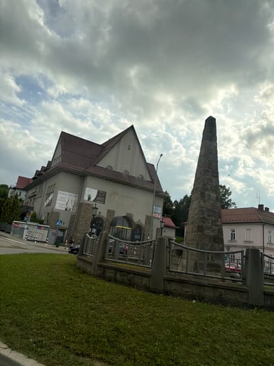 The image depicts a view of a plaza in Limanowa, Poland, featuring a tall, stone obelisk monument in the foreground. The monument is surrounded by a low, stone wall with a metal railing. To the left of the monument is a building with a distinctive, steeply pitched, reddish-brown roof and a light-colored facade. This building has several windows and appears to house businesses, as indicated by signage. One sign reads "Ślubny Zakątek," suggesting a bridal shop. Further to the left, a street with parked cars and a white structure that might be a car wash or vending machine is visible. The background shows more buildings with reddish roofs and trees, under a cloudy sky. The overall scene appears to be a public space in a town or city.