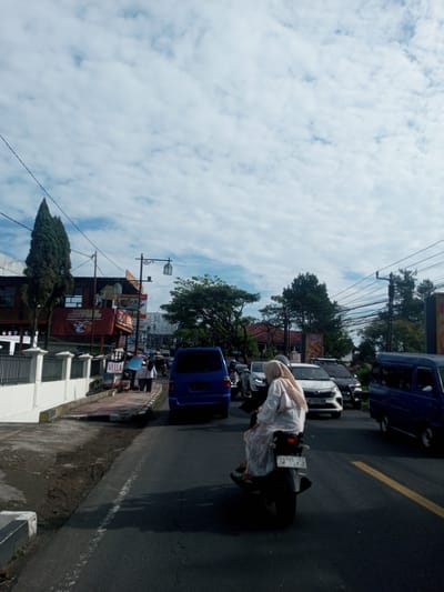 A street scene shows traffic, including a blue minivan and a white car, on a road marked with a yellow line. In the foreground, two individuals on a motorcycle are driving toward the camera. The person in the back is wearing a light-colored hijab and dress. The environment includes buildings, trees, power lines, and a sky with scattered clouds. The image appears to be taken in an urban area. There is no evidence to support the location being Zeyathiri District, Myanmar.