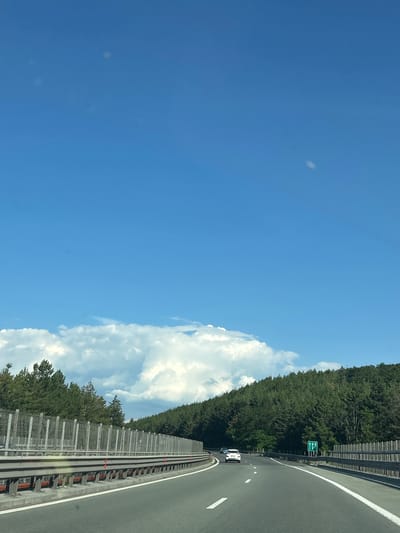 The media file captures a view from a vehicle traveling on a highway in Novi Han, Bulgaria. The scene is dominated by a bright blue sky with scattered white clouds. On the right side of the frame, a dense forest lines the highway, with the trees appearing lush and green, suggesting a time during the warmer months. The highway itself is paved and has white lane markings. A modern sound barrier runs along the side of the road, separating the highway from the forest.

Several vehicles are visible on the highway, including a white car and a smaller blue car, indicating ongoing traffic. A green road sign with white symbols is also present, directing traffic, though the specific directions are not clearly legible. The perspective suggests the image was taken from inside a moving vehicle, looking out towards the road and the surrounding landscape. The bright sunlight and clear sky imply it is daytime, likely mid-morning or afternoon. The overall atmosphere is one of travel and clear weather. There is no visible text that can be read clearly.