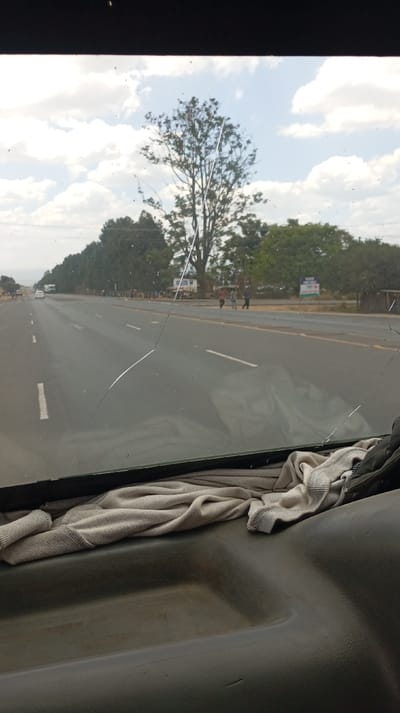 The image provides a view from inside a vehicle, looking out through its windshield onto a road and the surrounding landscape during daytime.

In the foreground, the dark grey or black dashboard of the vehicle is visible, with a light-colored, possibly beige or light brown, cloth or towel crumpled and resting on it, extending across the lower edge of the windshield.

The windshield itself is a prominent feature, appearing somewhat dirty or dusty, and notably cracked. A major crack originates from the top center-left, extends diagonally downwards and to the left, then sharply bends towards the bottom right corner, traversing a significant portion of the glass. Other smaller cracks are visible, including one near the top right and another near the bottom right. The reflections of the dashboard and the cloth are subtly visible on the inner surface of the windshield.

Beyond the windshield, a wide, paved multi-lane road stretches into the distance, suggesting a highway or a main thoroughfare. White dashed lines mark the lanes, and solid yellow lines are visible along the road's edges. In the far distance on the left lane, a white vehicle, possibly a truck or bus, can be seen.

The road is flanked by trees on both sides, with a denser line of trees on the left side extending into the horizon. On the right side of the road, several people are walking on what appears to be a dirt or unpaved shoulder parallel to the road. They are in the mid-distance and appear to be casually strolling.

Further beyond the people on the right, a blue and white sign is partially visible. While difficult to read definitively, it appears to have white text on a blue background, possibly indicating "PLOTS FOR SALE" or similar commercial information. A large, prominent tree with sparse branches stands centrally in the upper background, silhouetted against the sky.

The sky is bright and partly cloudy, with scattered white cumulus clouds against a lighter blue background, indicating a clear, sunny day. The overall impression is of a journey through a semi-rural or suburban area.