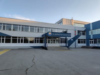 A school building is pictured under a clear blue sky with a few wispy clouds, suggesting it is daytime. The building is modern, with a gray facade and large windows. A blue and white geometric design adorns the lower part of the building. The entrance features a set of stairs leading to double glass doors, flanked by railings. Above the entrance, a blue awning displays a crest and text in Cyrillic script that reads "СУ "ДимИТЪР БЛАГОЕВ"". A Bulgarian flag is prominently displayed on a flagpole to the right of the entrance. The foreground shows a paved area, likely a courtyard or parking lot, with a yellow line marking an area. The overall impression is of a functional and well-maintained educational institution.