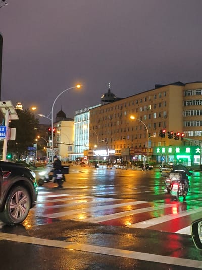 This image depicts a wet city intersection in Jixi, China, during nighttime. The wet asphalt street reflects the colorful lights from traffic signals, streetlights, and neon signs on the surrounding buildings. A pedestrian crossing, marked with red and white stripes, is visible in the foreground, slick with rain. On the street, two individuals are riding scooters; one in the foreground is facing away from the camera, and another is further back, also moving away. A black car is parked on the left side of the frame. The buildings in the background are illuminated, some with windows lit from within. The sky is overcast and dark, typical of a rainy night.