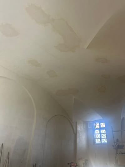 This is a close-up, eye-level, interior shot of a ceiling in what appears to be a historic building undergoing renovation, possibly located in Rosice, Czechia. The ceiling is made of a pale, slightly textured material and shows numerous splotchy water stains in various shades of beige and light brown, suggesting previous water damage. These stains are unevenly distributed across the arched sections of the ceiling. In the background, a window with a diamond-shaped grille is visible, and a portion of a scaffolding structure is present on the right side of the frame. The lighting is somewhat diffuse, with a bright glare emanating from the window area. The overall impression is one of an unfinished space with visible signs of repair and restoration work.