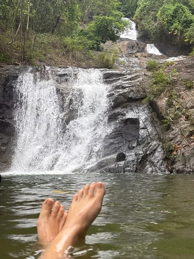 The image shows a waterfall in Thai Mueang, Thailand. The waterfall cascades down a rocky cliff face covered in lush green vegetation. In the foreground, a pair of human feet are submerged in the water. The water is clear and reflects the surrounding environment.
