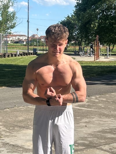 The image shows a muscular, shirtless man posing outdoors, with his biceps flexed. He wears white athletic shorts and a watch on his left wrist. The background includes a park-like area with trees and outdoor exercise equipment, suggesting a public space. The city's architecture is visible in the distance, indicating an urban location.