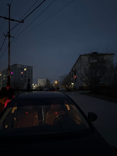 The image shows a nighttime street scene in Zhatay, Russia. Buildings line the street, lit by streetlights and a few interior lights. A vehicle is parked on the street, obscuring some of the view, with objects visible inside the car. Electrical wires are visible in the sky. A statue is visible, potentially of Lenin or another figure associated with the Soviet era.