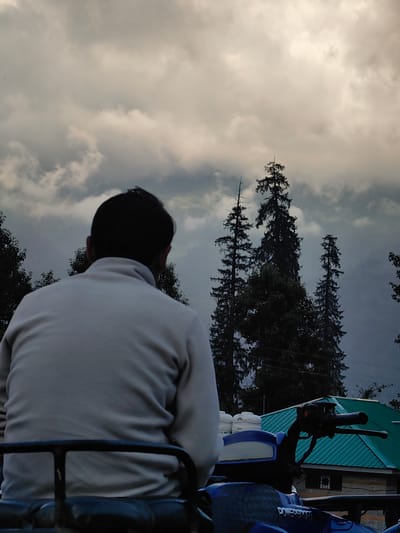 An individual, viewed from behind, sits on an ATV in Solang, India, facing a misty, cloud-covered mountain landscape. Tall, dark green coniferous trees punctuate the scene, reaching towards a sky filled with dramatic, overcast clouds. In the foreground, a blue ATV is partially visible, with the person's light-colored sweater and dark hair discernible. A green-roofed building with a brick base is situated behind the ATV, adding a touch of human habitation to the rugged natural setting. The overall atmosphere is somber and serene, evoking a sense of solitude amidst the grandeur of nature.