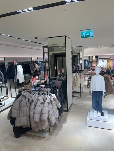 The image displays the interior of a brightly lit, modern clothing store, likely a fashion retailer or department store, featuring a clean design with light-colored tiled floors and white ceilings. The environment appears spacious and well-organized.
In the upper part of the frame, a mannequin is prominently displayed, wearing a plain white long-sleeved top and blue jeans, standing on a white pedestal. Another mannequin's torso, adorned with light pink and yellow tops, is partially visible in the background on the right. The store utilizes black-framed clothing racks and display tables to showcase merchandise.
A large quantity of light-colored (beige or light grey) puffer jackets or coats is neatly stacked on a display table in the foreground and mid-ground on the right side of the image. Adjacent to these, various other clothing items, including patterned tops, darker garments, and lighter casual wear, are arranged on hangers on racks and folded on tables.
Several red and white price tags are visible on the clothing displays. Notable text on these tags includes "TOUT À 79" (Everything for 79), "TOUT À 35" (Everything for 35), "TOUT À 25" (Everything for 25), and "TOUT À 17" (Everything for 17), suggesting a sale or fixed-price offering. Given the location context of Geneva, Switzerland, these prices are likely in Swiss Francs (CHF).
Mirrored panels, possibly integrated into structural columns or display units, reflect sections of the store, contributing to the bright and expansive atmosphere. High on a wall or ceiling, a green illuminated exit sign is visible, depicting a running person symbol and an upward-pointing arrow. The scene captures a typical retail setting during what appears to be regular business hours, with no actual customers or staff visible.