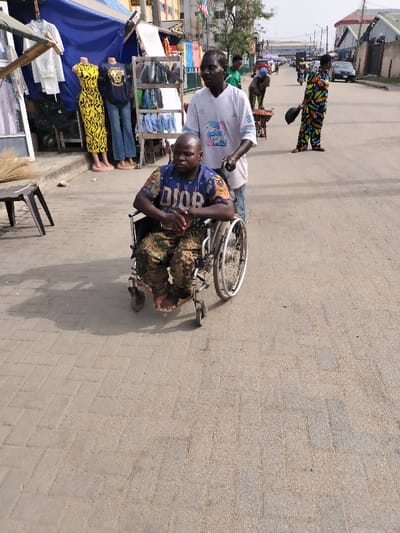 A daytime street scene in Alaiyabiagba, Nigeria, depicts a dark-skinned male in a blue "DIOR" t-shirt and camouflage trousers seated in a manual wheelchair. His bare feet rest on the footrests, and his hands are clasped in front. Another dark-skinned male, wearing a white t-shirt with a blue graphic, stands behind him, pushing the wheelchair along a light-colored paved sidewalk. To their immediate left, a shopfront displays clothing on mannequins, including a yellow animal-print maxi dress. The asphalt road to the right features pedestrians and vehicles in the mid-ground and distance, including a person in a colorful patterned jumpsuit holding a dark object and another pushing a handcart. Commercial buildings line the street.