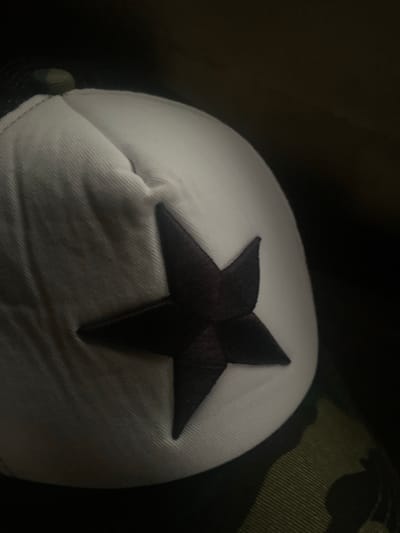 A close-up, dark, and slightly blurry shot shows a portion of a white baseball cap with a dark four-pointed star emblem. The cap is angled, with the brim and the front panel visible. The background is dark and indistinct. The bottom right of the image shows a hint of a camouflage pattern on what appears to be fabric, possibly part of the cap or clothing. The scene is indoors, with minimal lighting. No people, actions, or interactions are discernible. The location in Tavo, Nigeria, cannot be determined from the visual information provided.